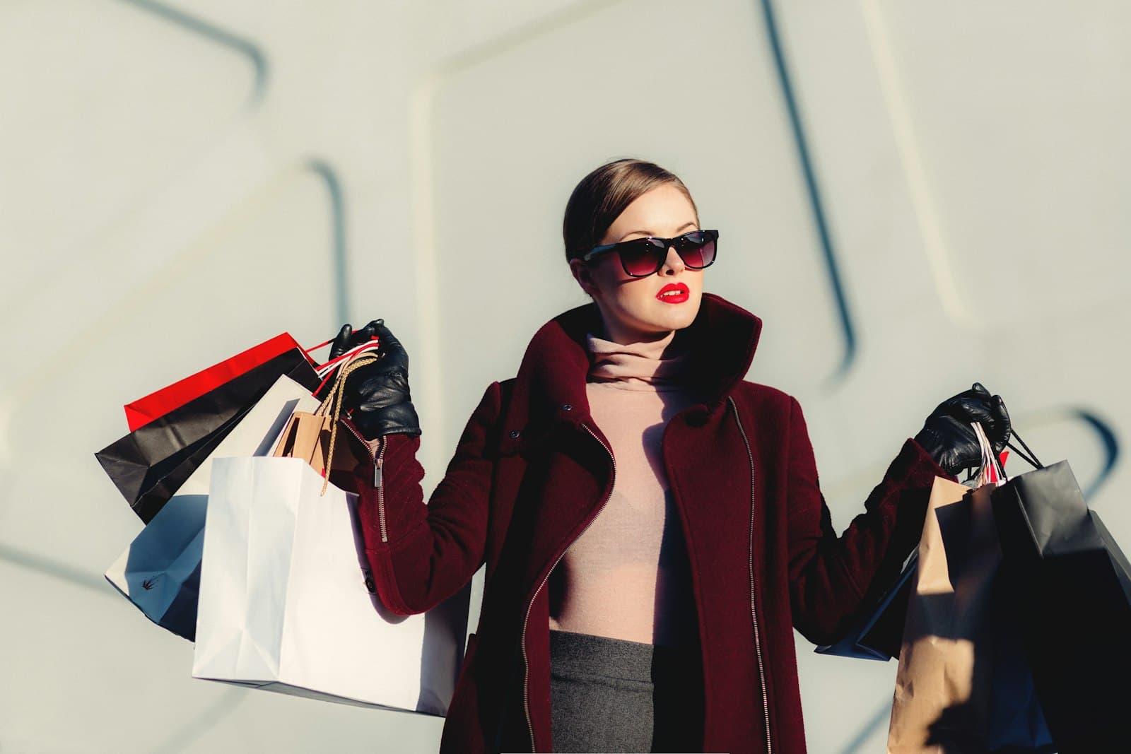 A woman in a burgundy coat and sunglasses carrying shopping bags against a pale wall.