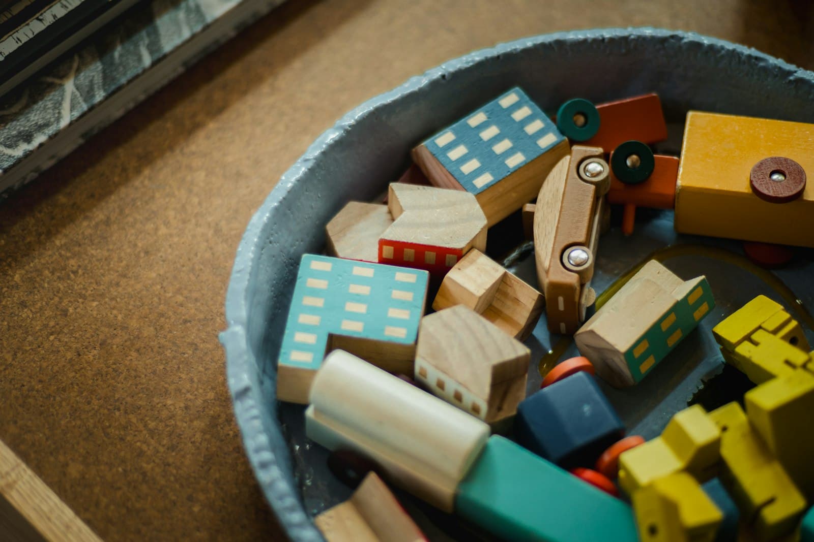 A ceramic bowl of colorful wooden toy houses, cars, and blocks on a cork table.